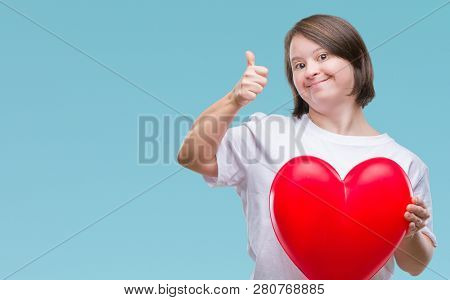 Young adult woman with down syndrome holding red heart over isolated background happy with big smile doing ok sign, thumb up with fingers, excellent sign