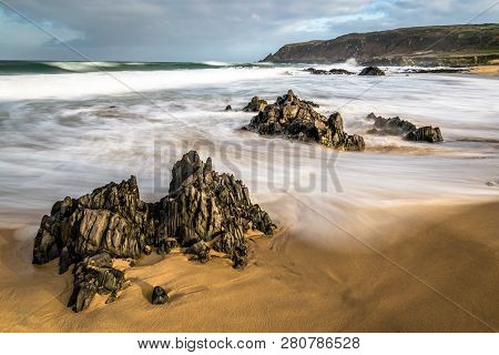Blurred Waves Swirling Around Rocks On A Sandy Beach In Ireland.