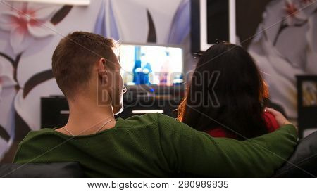 Young Cute Couple Sitting On A Couch Watching Tv Together. Young Man Hugs His Girlfriend