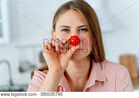 Beautiful Cheerful Person Holds Cherry Tomato Near Nose. Ktchen Blurred Background. Funny Facial Exp
