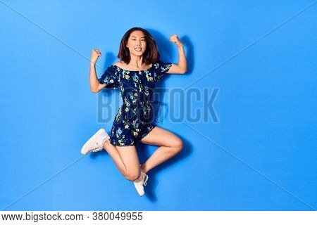 Young beautiful chinese woman wearing casual clothes smiling happy. Jumping with smile on face doing strong geture over isolated blue background