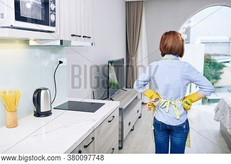 Housewife In Apron And Rubber Gloves Looking At Apartment She Just Cleaned, View From The Back