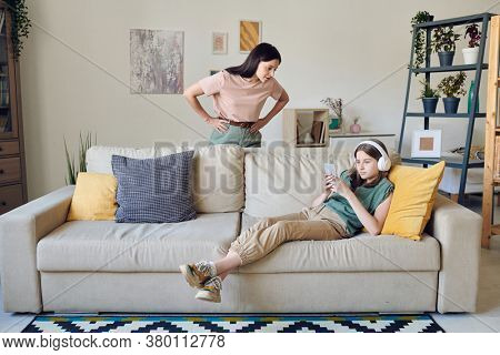 Strict mother keeping her hands on waist while standing by couch where her lazy teenage daughter with smartphone listening to music