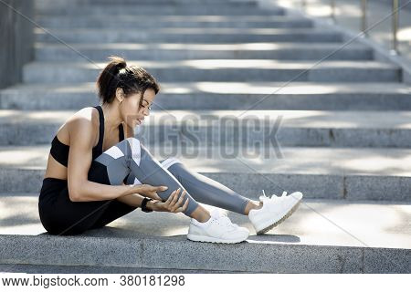 Upset African Woman In Sport Clothes Sitting On Stairs, Massaging Ancle, Side View, Empty Space