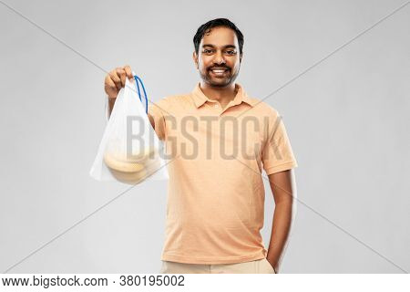 food, sustainability and eco living concept - happy smiling young indian man in polo t-shirt holding reusable string bag with bananas over grey background