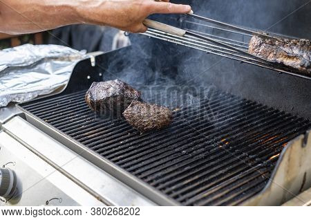 Man Cooking Beef On The Grill Outside, Flipping The Meat With Tongs.