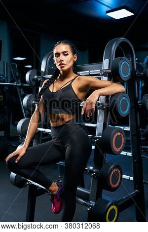 Side Portrait Of Female Bodybuilder With Long Brunette Braids Posing Near Stand With Sports Equipmen
