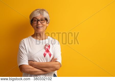 Senior woman on color yellow background. Pink ribbon like a symbol of breast cancer awareness. Support people living with tumor illness.