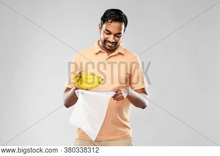 food, sustainability and eco living concept - happy smiling young indian man in polo t-shirt putting bananas into reusable string bag over grey background