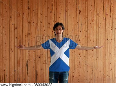 Man Wearing Scotland Flag Color Shirt And Standing With Arms Wide Open On The Wooden Wall Background
