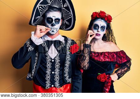 Young couple wearing mexican day of the dead costume over yellow mouth and lips shut as zip with fingers. secret and silent, taboo talking 