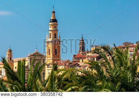 Bell tower of Basilica of Saint Michel Archange under blue sky as seen through the palms in old town of Menton, France.