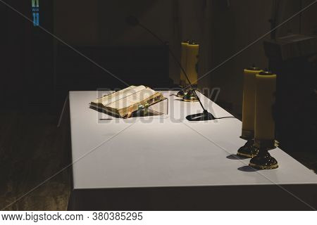 Dark Church Altar With Open Bible Book And Yellow Candles On Table With White Tablecloth
