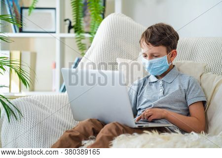 Focused Boy In A Mask, Sitting On The Sofa With A Laptop During The Quarantine Period. He Does Not G