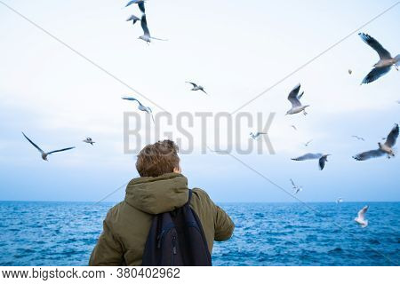 A Young Guy In A Green Jacket Stands With His Back And Feeds Seagulls On The Seashore