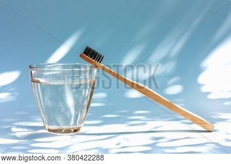 Bamboo Toothbrush And Glass Of Water On Light Blue Pastel Background With Shadows Of Natural Light. 