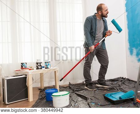 Guy Singing During Home Repair Using Roller Brush Dipped In Blue Paint. Hipster Renovating His House