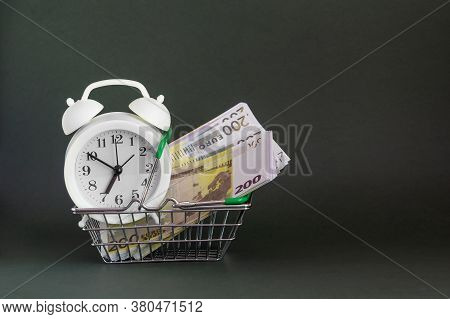 A White Alarm Clock And A Wad Of Paper Money, In A Shopping Basket, On A Dark Isolated Background. T