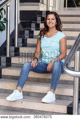 Tanned Young Women Wearing T-shirt And Jeans Sits On The Steps And Smiling