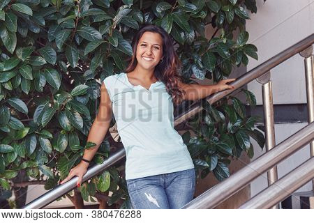 Tanned Young Women Wearing T-shirt And Jeans Stays On The Stairs Near The Plant And Smiling