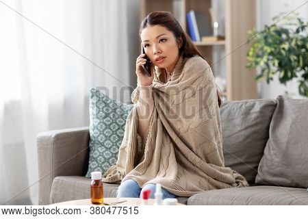 technology, health and cold concept - sick asian woman in blanket calling on smartphone at home