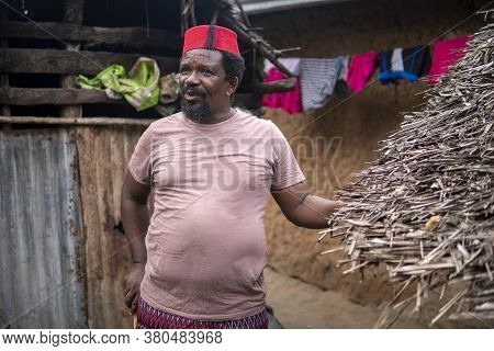 An African Older Man In Red Muslim Taqiyyah Fez Hat Posing With A Stick For Lame People On Yard Near