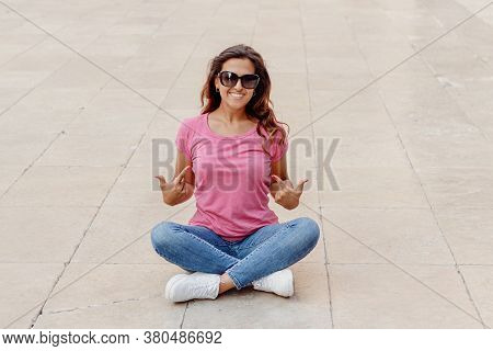 Young Women Wearing T-shirt And Jeans Sits On The Pavement And Shows In The  Center