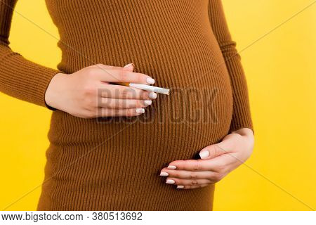 Close Up Of Pregnant Woman In Brown Dress With A Cigarette Against Her Belly At Yellow Background. D