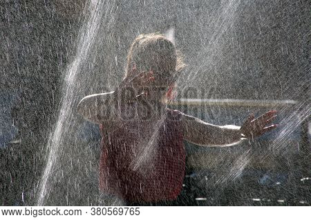 A Girl Bathes In A Fountain On A Sunny Day. Heat, Spray Drops From The Fountain Refresh A Young Girl