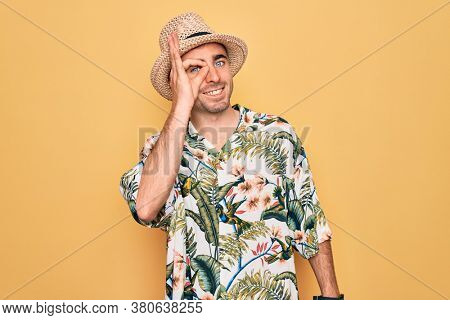 Young handsome man with blue eyes on vacation wearing summer florar shirt and hat doing ok gesture with hand smiling, eye looking through fingers with happy face.
