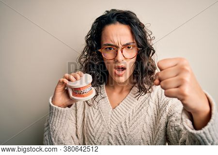 Young beautiful woman with curly hair holding plastic teeth with dental baces annoyed and frustrated shouting with anger, crazy and yelling with raised hand, anger concept