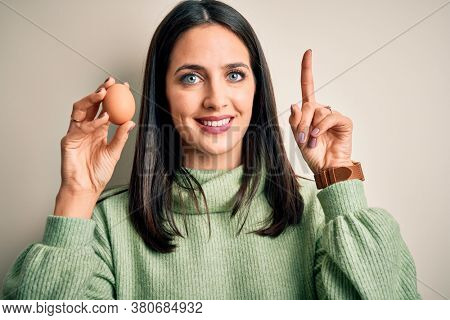 Young brunette woman with blue eyes holding fresh raw egg over isolated background surprised with an idea or question pointing finger with happy face, number one