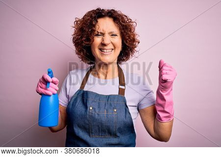 Middle age curly hair woman cleaning doing housework wearing apron and gloves using spayer screaming proud and celebrating victory and success very excited, cheering emotion