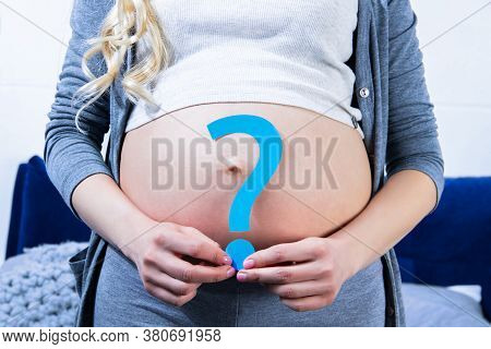 Closeup Of Big Pregnant Female Belly. Young Woman Is Holding Blue Paper Question Sign Near Abdomen. 