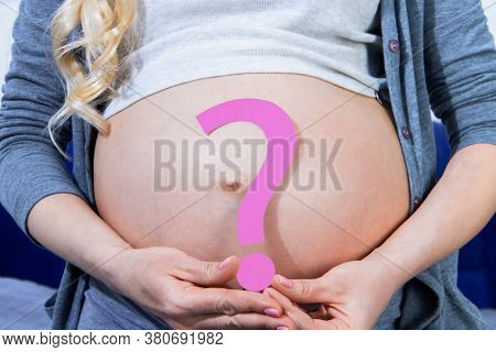 Closeup Of Big Pregnant Female Belly. Young Woman Is Holding Pink Paper Question Sign Near Abdomen. 