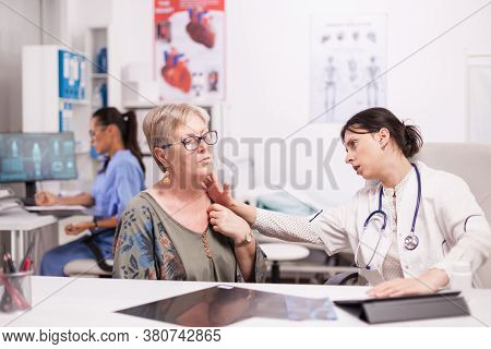 Doctor In Hospital Cabinet Examining Neck Glands Of Senior Patient. Mature Woman With Thyroid Discom