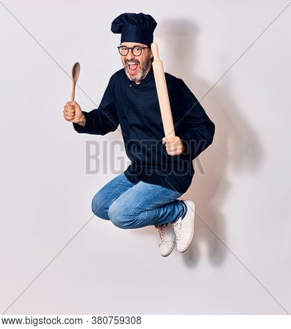 Middle age handsome hispanic man wearing glasses and cook uniform smiling happy. Jumping with smile on face holding kneader and spoon over isolated white background