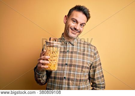 Young handsome man holding jar with dry italian pasta macaroni over yellow background with a happy face standing and smiling with a confident smile showing teeth