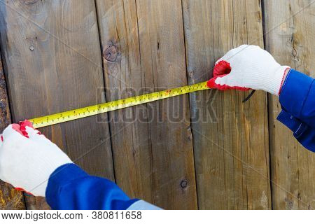 Male Hands In Work Gloves With A Yellow Tape Measure Against A Background Of Dark Boards Measure The
