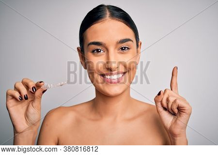Young beautiful brunette woman holding dental aligner orthodontic to teeth corretion surprised with an idea or question pointing finger with happy face, number one