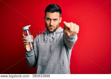 Young handsome sportsman drinking bottle of water to refeshment over red background pointing with finger to the camera and to you, hand sign, positive and confident gesture from the front
