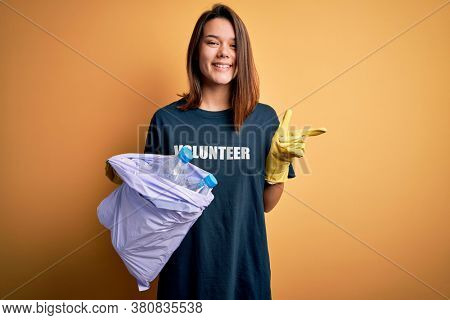Beautiful volunteer girl caring environment doing volunteering holding bag with rubish bottles very happy pointing with hand and finger to the side