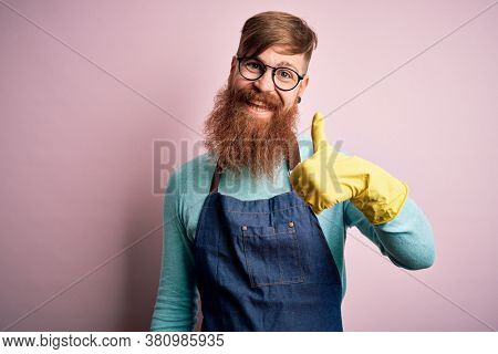 Irish redhead housekeeping man with beard wearing apron and washing gloves doing happy thumbs up gesture with hand. Approving expression looking at the camera showing success.