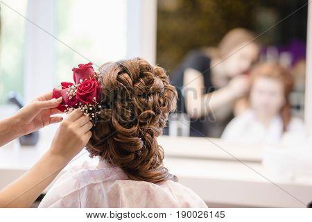 Female hairdresser standing and making hairstyle to cute lovely young woman in beauty salon. Hairstyle with red roses.