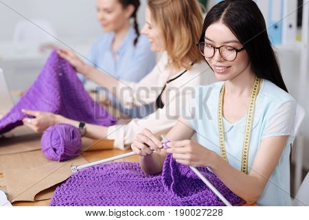 Handicraft time. Beautiful young women sitting at the table in the atelier and knitting scarves with purple threads using big knitting needles.