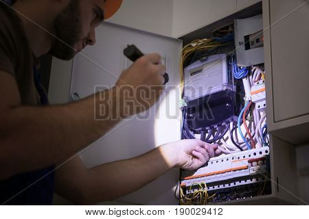 Young electrician with flashlight near switch box in dark room