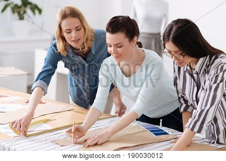 Upsurge of creativity. Two young brunette female tailors tracing patterns on fabrics with pencils while their colleague measuring the width of fabrics
