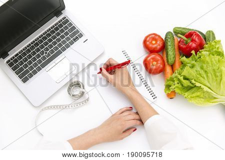 Vegetable diet nutrition or medicaments concept. Doctors hands writing diet plan ripe vegetable composition laptop and measuring tape on white background