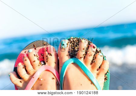 Funny woman feet with multi-colored pedicure in slippers on summer pebble beach background. Inspirational vibrant outdoors horizontal close-up image.