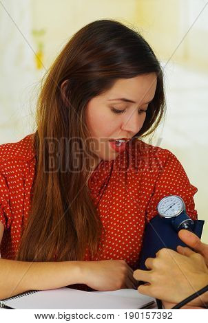 Beautiful young patient using a manual tensiometer in her arm, in a doctor consulting room background.